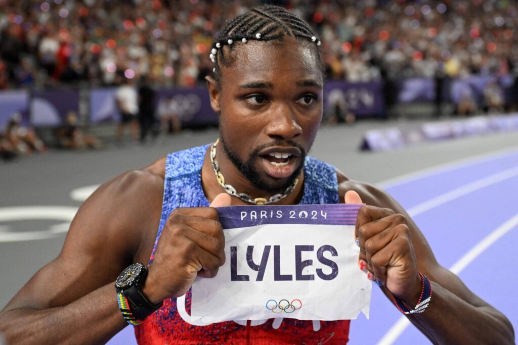 Noah Nyles comemora na pista de atletismo mostrando o papel com o seu nome em destaque, “Lyles”, e, em letra menor, no alto do papel, “Paris 2024”.