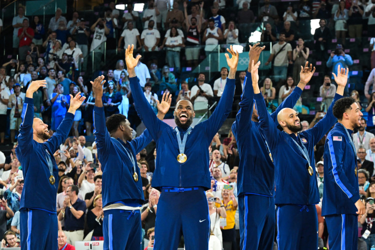 Jogadores da seleção de basquete dos Estados Unidos comemoram, no meio da quadra, com os braços estendidos e a medalha de ouro no peito, sob os aplausos da plateia.